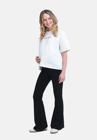 Vit bomullst-shirt med texten "COOL MAMA", matchad med högmidjade svarta utsvängda byxor. Modellen står avslappnat med en hand på sin mage.