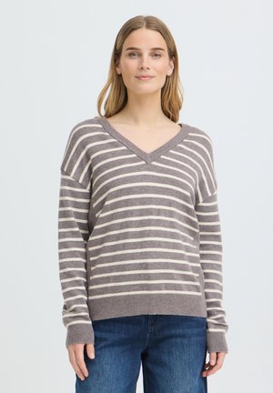 Femme aux cheveux lisses châtain clair portant un pull rayé gris et blanc à col en V et un jean bleu devant un fond clair uni.