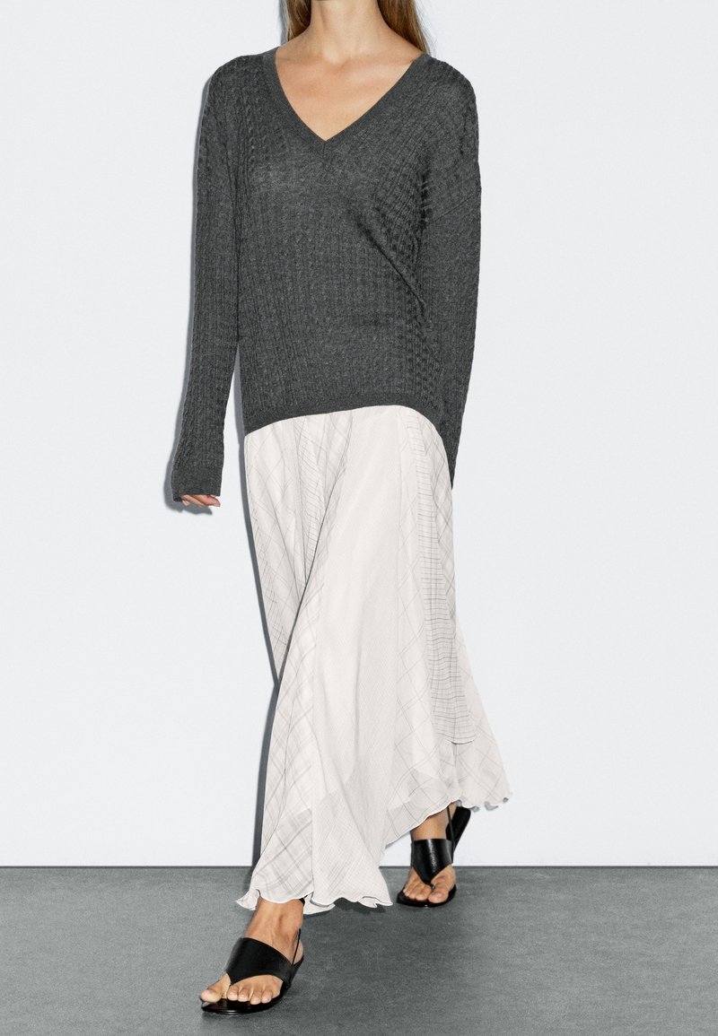 Femme portant un pull en maille torsadée gris foncé à col en V, une jupe blanche à motifs longueur cheville et des sandales à talons ouvertes noires.