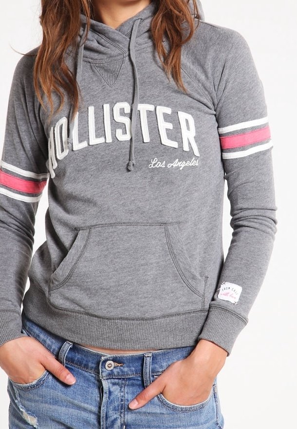 Donna che indossa una felpa con cappuccio grigia Hollister con strisce rosa e bianche sulle maniche e jeans blu strappati, con le mani nelle tasche.