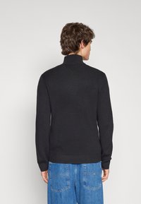 Maglione nero a costine con colletto alto e maniche lunghe, caratterizzato da un motivo a maglia testurizzato e un orlo aderente, indossato con jeans blu.