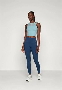 Young woman standing in blue sleeveless crop top, navy high-waist leggings, and white sneakers against plain background.