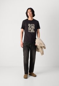 T-shirt nera con stampa di foglie, abbinata a jeans scuri e una giacca beige chiaro. Modello che indossa scarpe slip-on color sabbia. Design semplice e casual.