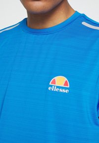 Modrá sportovní tričko vyrobené z texturované tkaniny s krátkými rukávy. Na hrudi se nachází barevné logo s červenými, žlutými a šedými akcenty.