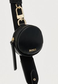 Kicsi, kerek, fekete bőr táska arany BOSS logóval, arany cipzárral, és egy csattal, amely egy széles fekete pánthoz van rögzítve.