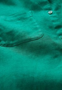 Camicia in lino verde con tasca frontale e dettagli in bottone, che presenta una texture liscia e una leggera lucentezza.