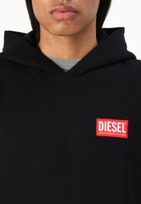 Sort bomulds hoodie med et stort rødt Diesel-logo-patch på brystet. Stoffet ser glat ud med en hætte med snoretræk og ribbede detaljer.