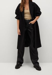 Manteau long noir avec un col structuré, porté sur un haut en maille beige clair et un pantalon large en similicuir noir. Chaussures épaisses noires.