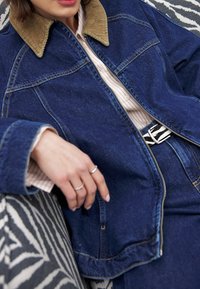 Denimu jaka ar korduroja apkakli, tumši zilā krāsā, ar rāvējslēdzēju, un redzamām šuvēm. Roka atpūšas uz svītrainā jostas un rakstainas virsmas.