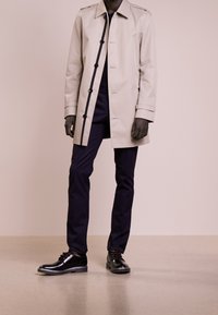 Impermeabile beige con bottoni neri, abbinato a pantaloni blu navy e scarpe in pelle nera, su uno sfondo neutro.