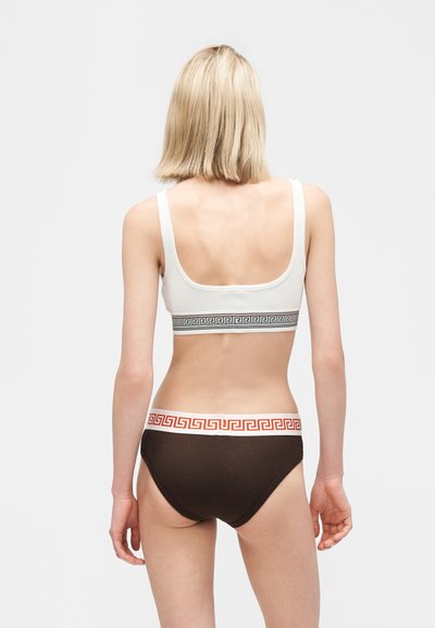 Γυναίκα, ορατή από πίσω, που φοράει λευκό αθλητικό σουτιέν με σχέδιο και σκούρα εσώρουχα με αντίθετη ζώνη με σχέδια σε γκρι φόντο.