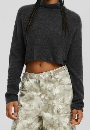 Personne portant un pull court gris foncé et un pantalon cargo tie-dye gris clair avec des cheveux tressés tombant sur les épaules.