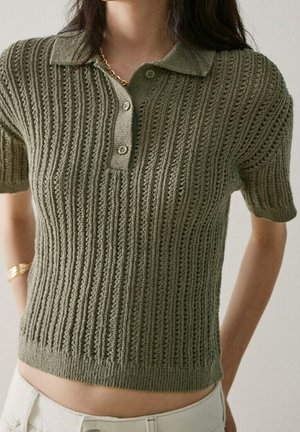 Femme portant un polo en maille à manches courtes vert olive avec un motif côtelé et un pantalon beige, accessoirisée de bijoux en or.