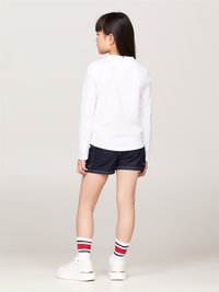 Camicia bianca a maniche lunghe con dettaglio a V sul retro, abbinata a shorts di jeans scuri. Indossa sneakers bianche e calze a righe con accenti rossi.