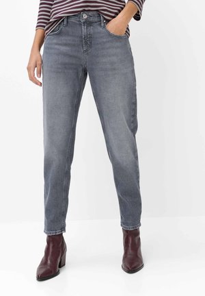 Person iført falmede grå jeans og mørkerøde ankelstøvler, hånd i lommen, står mod en ensartet hvid baggrund.