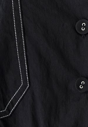 Melns audums ar baltu kontrastējošu diegu, kas izceļ kabatu, un divi melni pogas ar sudrabainiem acs caurumiem.