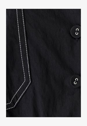 Melns audums ar baltu kontrastējošu diegu, kas izceļ kabatu, un divi melni pogas ar sudrabainiem acs caurumiem.