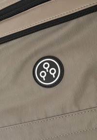 Harmaa kangas, jossa on hienovarainen rakenne, sisältää pyöreän mustan logopatchin, jossa on kolme reikää ja kontrastivalkoinen reunus. Musta vetoketju.