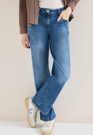 Person iført blå jeans med lige ben, hvide og lyseblå sneakers, brun jakke og hvid top, stående med den ene hånd i lommen.