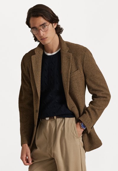 Rudas herringbone tipo švarkas su pritaikytu siluetu, derinamas su tamsiai mėlynu ripsu megztiniu ir šviesiai smėlio spalvos kelnėmis, demonstruojantis modernų sluoksniuotą įvaizdį.