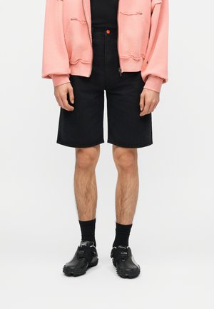 Person trägt eine rosa Jacke mit Reißverschluss, schwarze kniehohe Shorts, schwarze Socken und schwarze Sportschuhe und steht vor weißem Hintergrund.