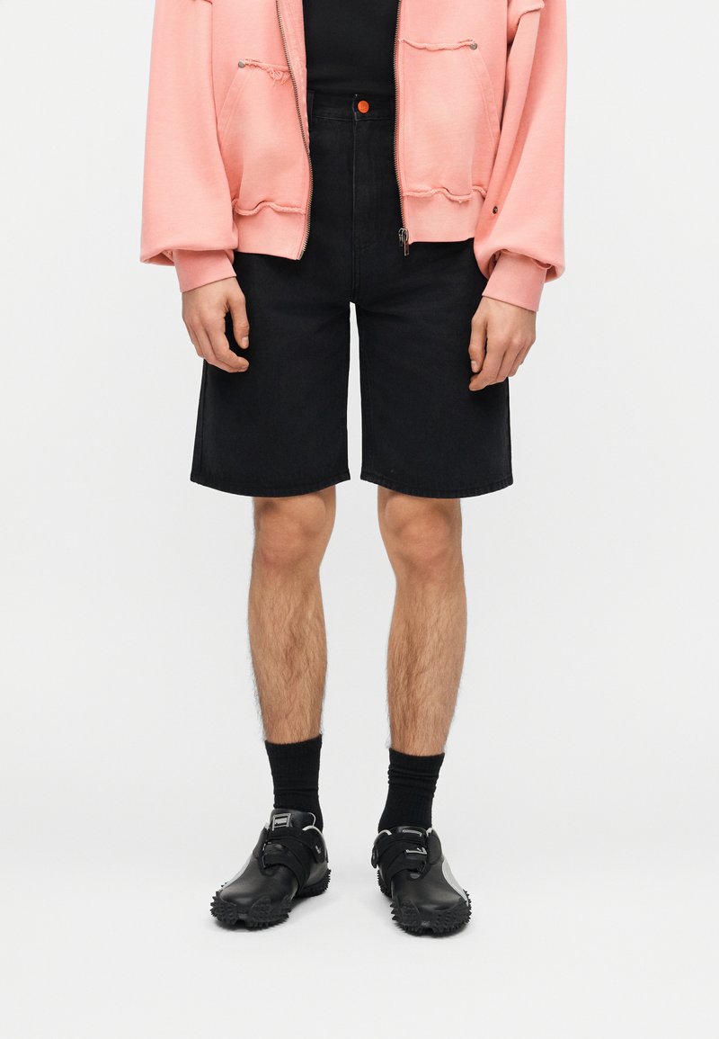 Άτομο που φορά ροζ ζακέτα με φερμουάρ, μαύρα σορτς μέχρι το γόνατο, μαύρες κάλτσες και μαύρα αθλητικά παπούτσια, στέκεται πάνω σε λευκό φόντο.