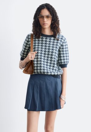 Kvinde med krøllet hår iført brune solbriller, en sort-hvid hundetandsmønstrede sweater, navy plisseret nederdel og holder en brun skuldertaske.