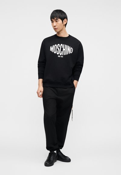 Μαύρο φούτερ με λογότυπο «MOSCHINO», σετ με μαύρο παντελόνι και μαύρα αθλητικά παπούτσια. Απλός σχεδιασμός και άνετη γραμμή. Λευκή επιγραφή ως διακοσμητικό στοιχείο.