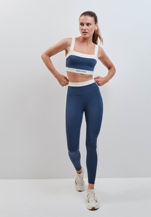 Vrouw in marineblauwe en witte sportbeha en legging die haar topje rechtzet terwijl ze staat tegen een effen lichtgrijze achtergrond.