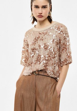 Vrouw draagt een beige kortemouwen top met pailletten en bruine hoge taille broek met een gestructureerde riem, poseert met handen in de zakken.