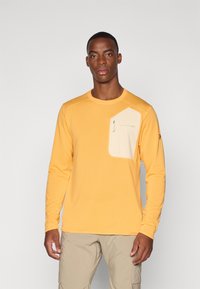 Langærmet, ribbet gul top med en cremfaret brystlomme, der har et logo og et let stof. Sammen med beige shorts.