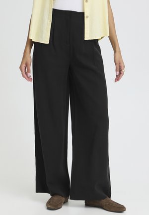 Personne portant un pantalon noir ample, des mocassins marron, un haut blanc et une chemise sans manches jaune boutonnée, debout devant un fond uni.