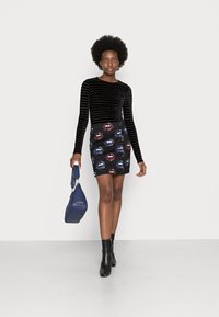 Blusa de veludo preto de mangas compridas combinada com uma saia mini estampada com lábios coloridos. A modelo leva uma mala azul-escura e usa botins pretos.