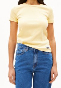 Žluté žebrované tričko s krátkým rukávem a kulatým výstřihem, spárované s světlomodrými vysokými džíny. Na lem trička je viditelná visačka.