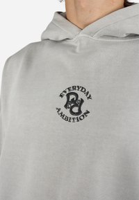 Lysegrå hætte sweatshirt lavet af blødt stof, med et frontgrafik der viser "EVERYDAY AMBITION" i sort og et rundt logo design.