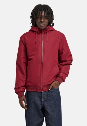 Personne avec des dreadlocks noires portant une veste rouge à capuche zippée et un jean bleu, debout avec une main dans la poche devant un fond uni.