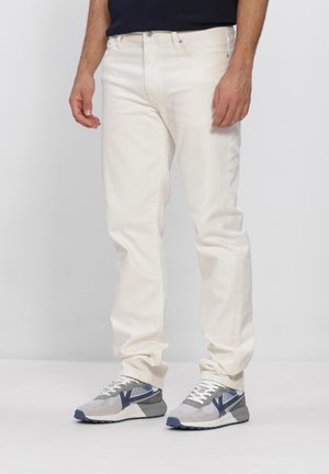 Mand iført off-white slim-fit jeans og grå sneakers med blå accenter, stående mod en ensfarvet lys baggrund.