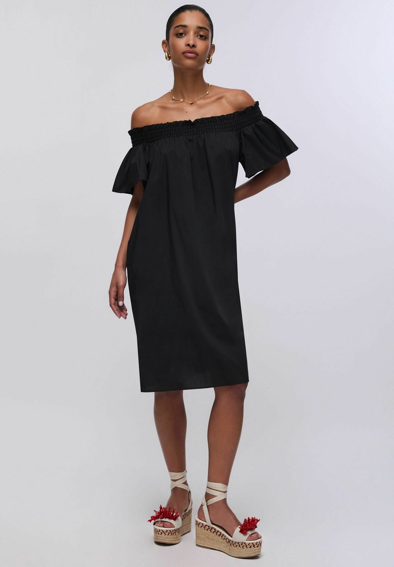 Femme portant une robe noire épaules dénudées longueur genou et des sandales compensées blanches avec des détails floraux rouges sur un fond clair.
