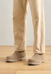 Botine din suede bej, cu panouri laterale elastice și talpă groasă din cauciuc, purtate cu pantaloni bej deschis, pe un podea din lemn.