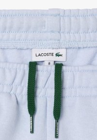 Shorts en sweat bleu clair avec une taille élastique, des cordons de serrage verts et une étiquette Lacoste affichant un logo de crocodile. Taille 8.