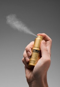 Guldsprayflaska hålls i en hand, märkt "STMNT." Svarta texturerade detaljer, fin dimma sprids från munstycket. Storlek: 0,14 oz (4 g).