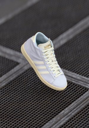 Biały sneaker Adidas z kremową podeszwą i sznurowadłami, trzema paskami po boku, unoszący się nad ciemną metalową powierzchnią z kratką.