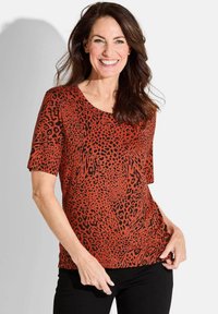 Rode top met een zwart dierenprintpatroon, korte mouwen en een ronde halslijn. Gemaakt van een gladde stof. Het model draagt het met donkere broek.