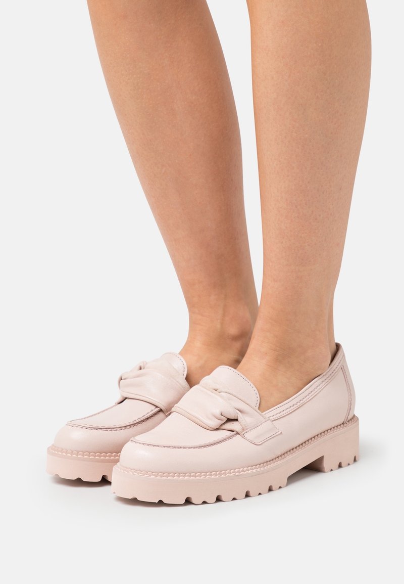 Mocassins en cuir rose clair avec semelles en caoutchouc épaisses et détail de bride torsadée portés pieds nus sur un fond uni.