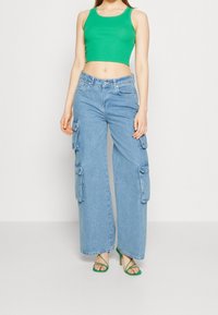 Jeans cargo de pierna ancha en azul claro con múltiples bolsillos y cintura alta, combinados con un top corto ajustado verde y tacones verdes con tiras.