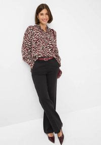 Rote und cremefarbene gemusterte Bluse mit langen Ärmeln, kombiniert mit schwarzen Hosen und einem passenden roten Gürtel. Glänzende burgunderfarbene Pumps vervollständigen den Look.