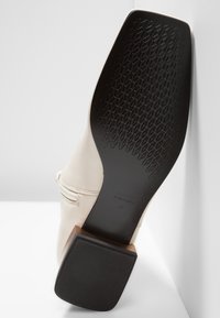Botte crème inclinée pour montrer la semelle noire avec motif texturé antidérapant et petit talon carré, posée contre un fond blanc.