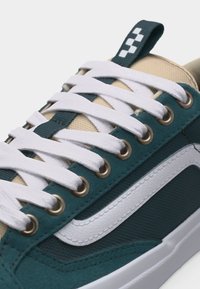 Κοντινό πλάνο ενός αθλητικού παπουτσιού σε τιρκουάζ και μπεζ χρώμα με λευκά κορδόνια περασμένα από μεταλλικά ροκανάκια και λευκή κυματιστή λεπτομέρεια στη πλευρική ρίγα.