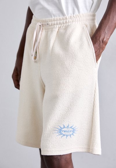 Beige strikkede shorts med elastisk midjebånd, snøring, sidelommer og et lyseblåt grafisk mønster på venstre ben.