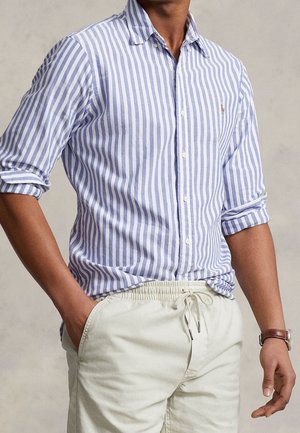Homme portant une chemise à manches longues rayée verticalement bleu et blanc, manches remontées, et un pantalon beige clair avec cordon, main dans la poche.